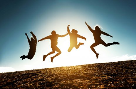 Four silhouettes of friends are jumping together against blue sky and sun